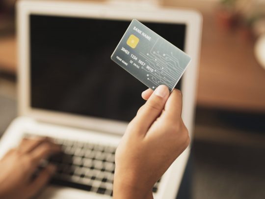 top-view-credit-card-held-in-hand-with-blurred-laptop-in-the-background
